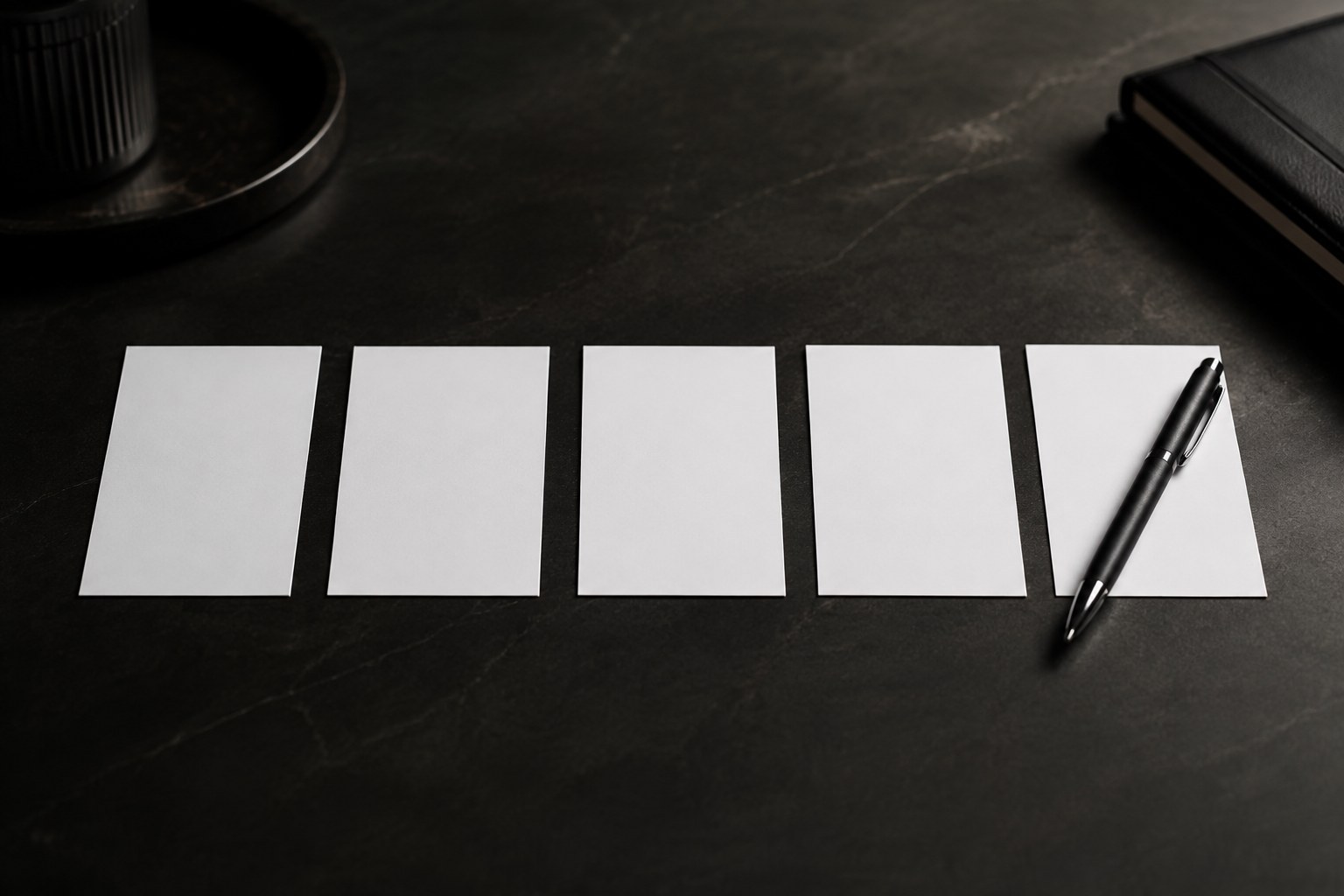 Five blank index cards in a row on a dark desk, a pen resting at the last card, no readable writing